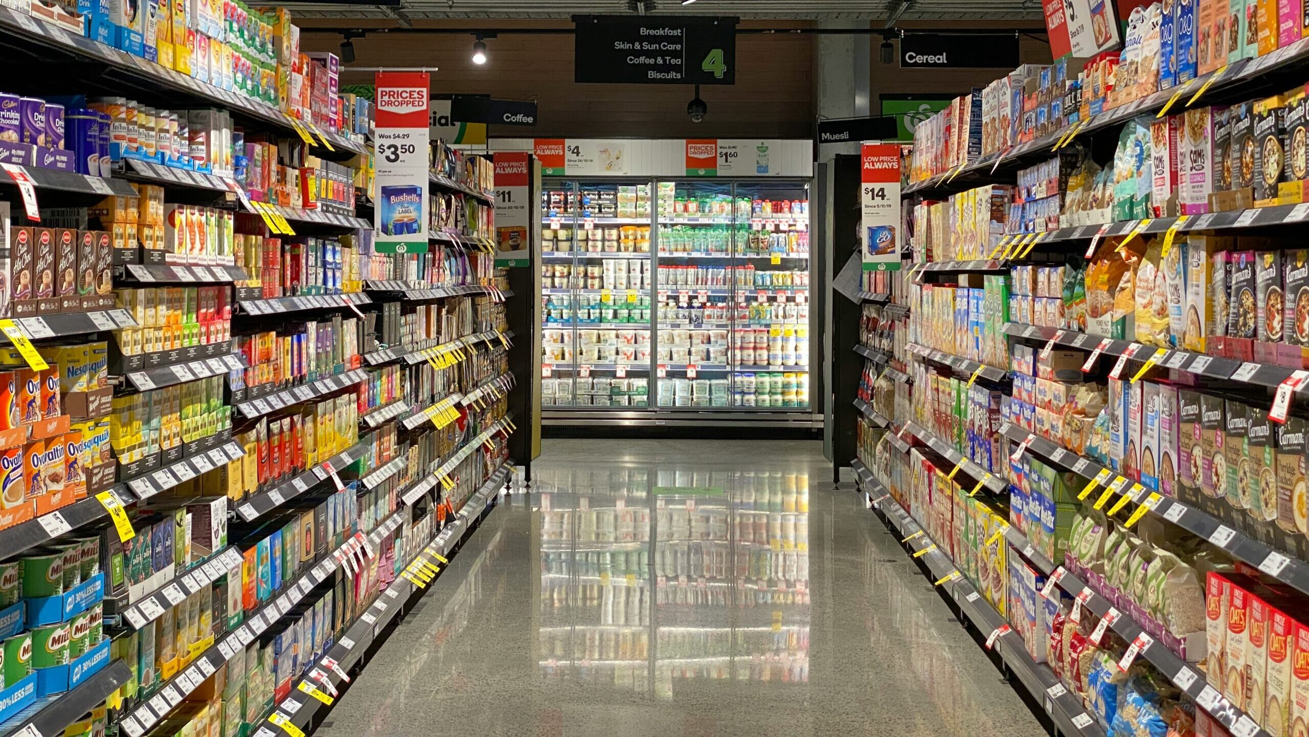 Two grocery store shelves containing boxed food products are shown on either side, leading to a refrigerator that contains chilled food products.