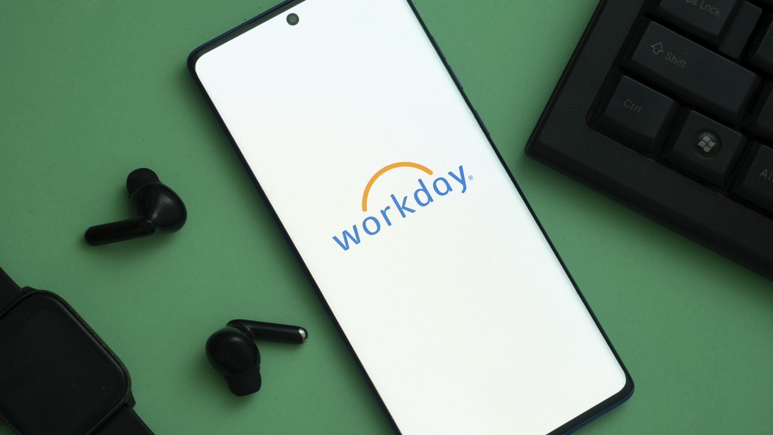 A smartphone shows the Workday logo, surrounded by earphones and a keyboard on a green background.