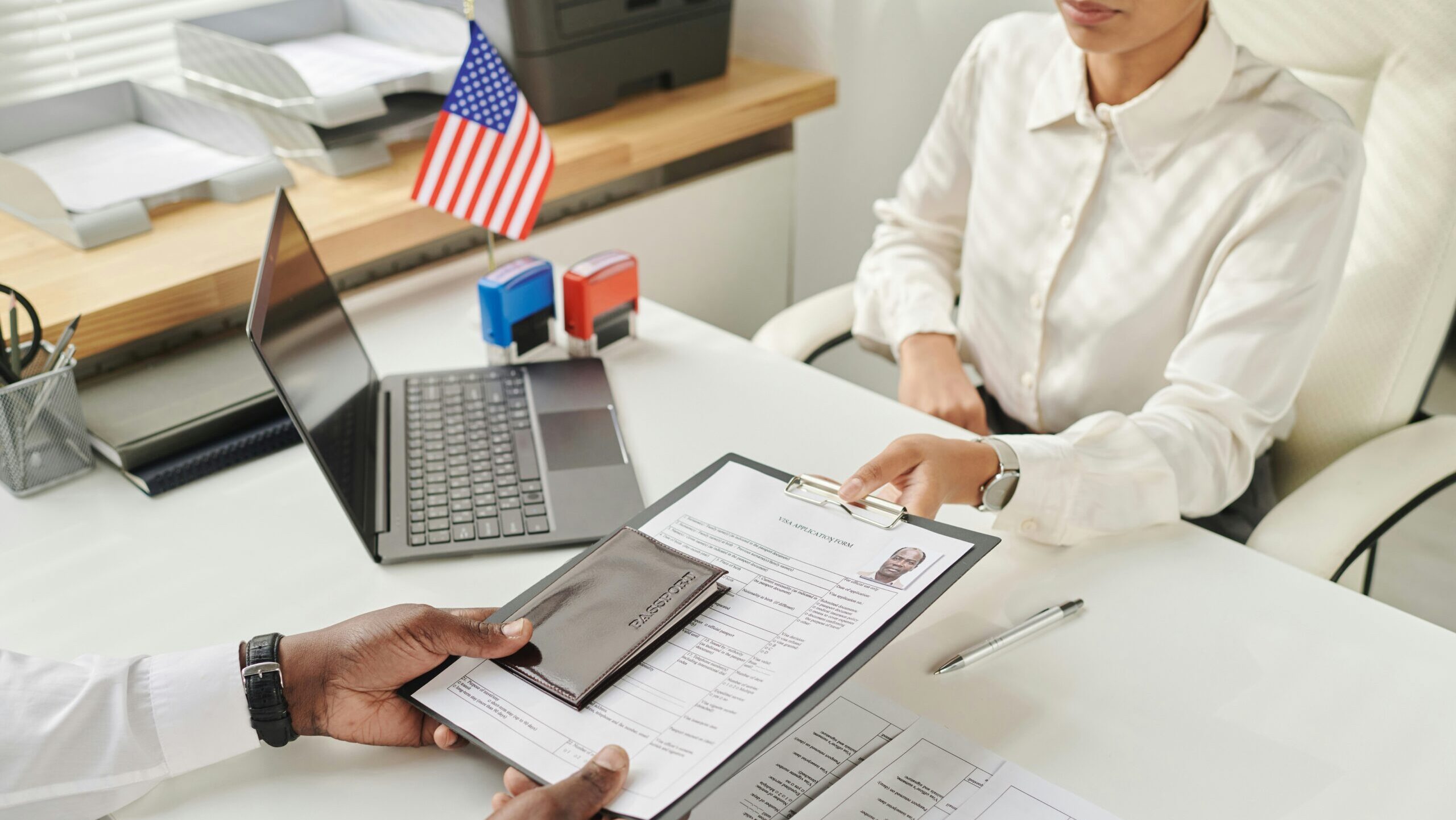 A person hands an immigration application for to a worker at a desk with an American flag on it.