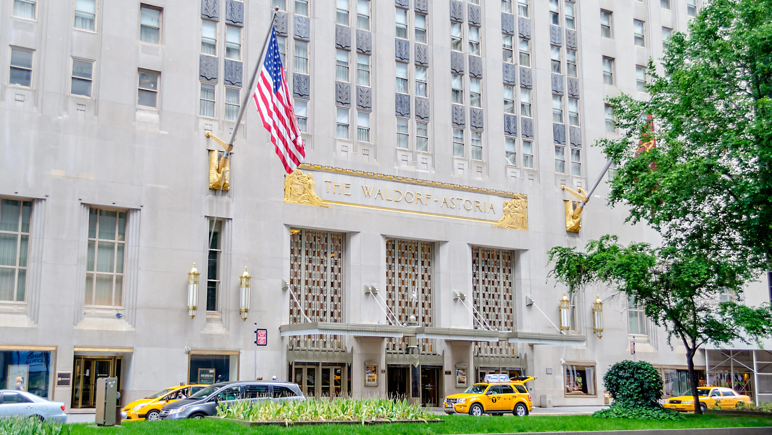 The exterior of the Waldorf Astoria Hotel in New York City is shown with cars and taxis passing by out front.