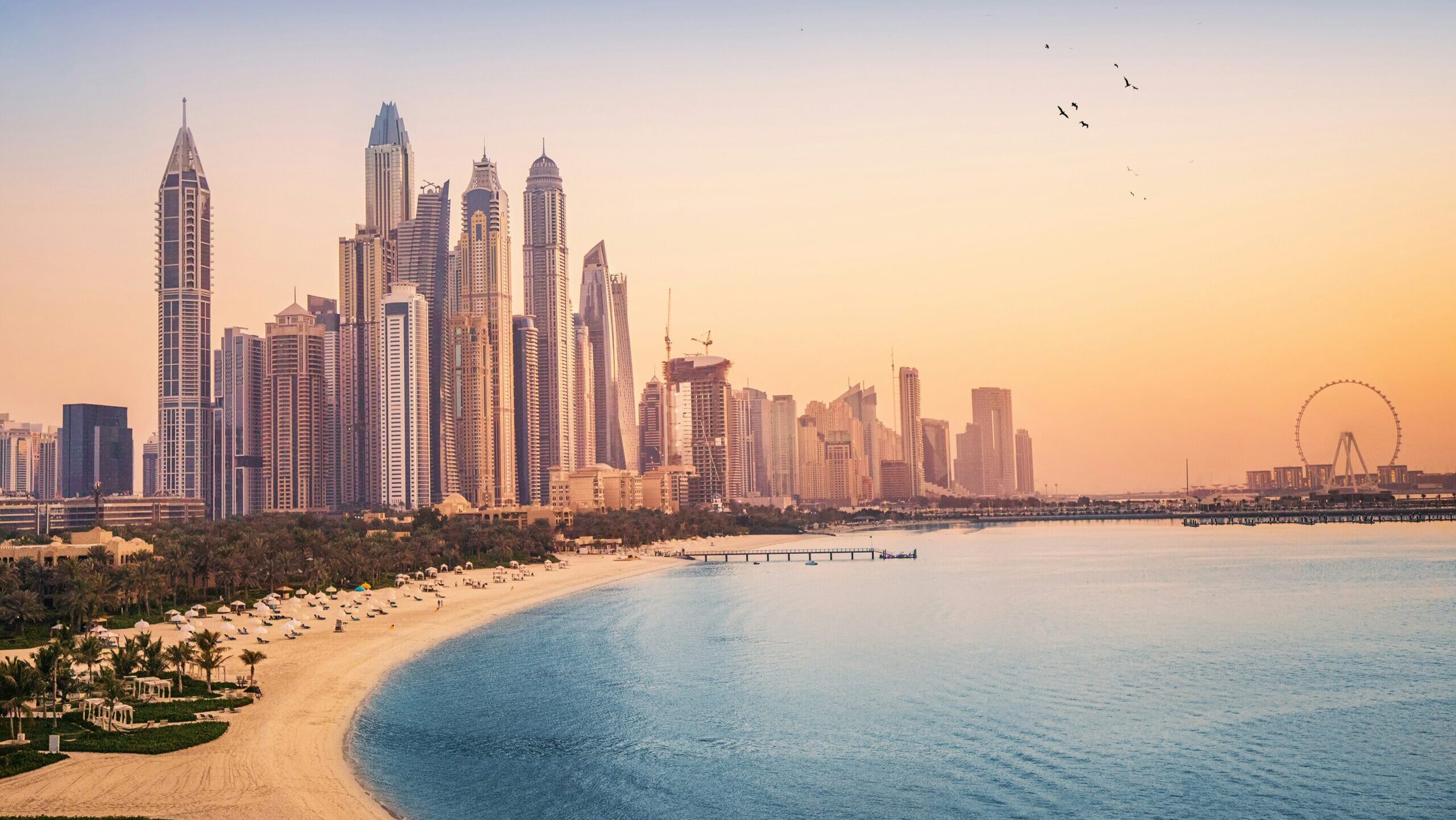 A sunset view of the Dubai Marina, including the city's Ferris wheel and the sandy beaches of the Persian Gulf, with the city skyline visible in the background.