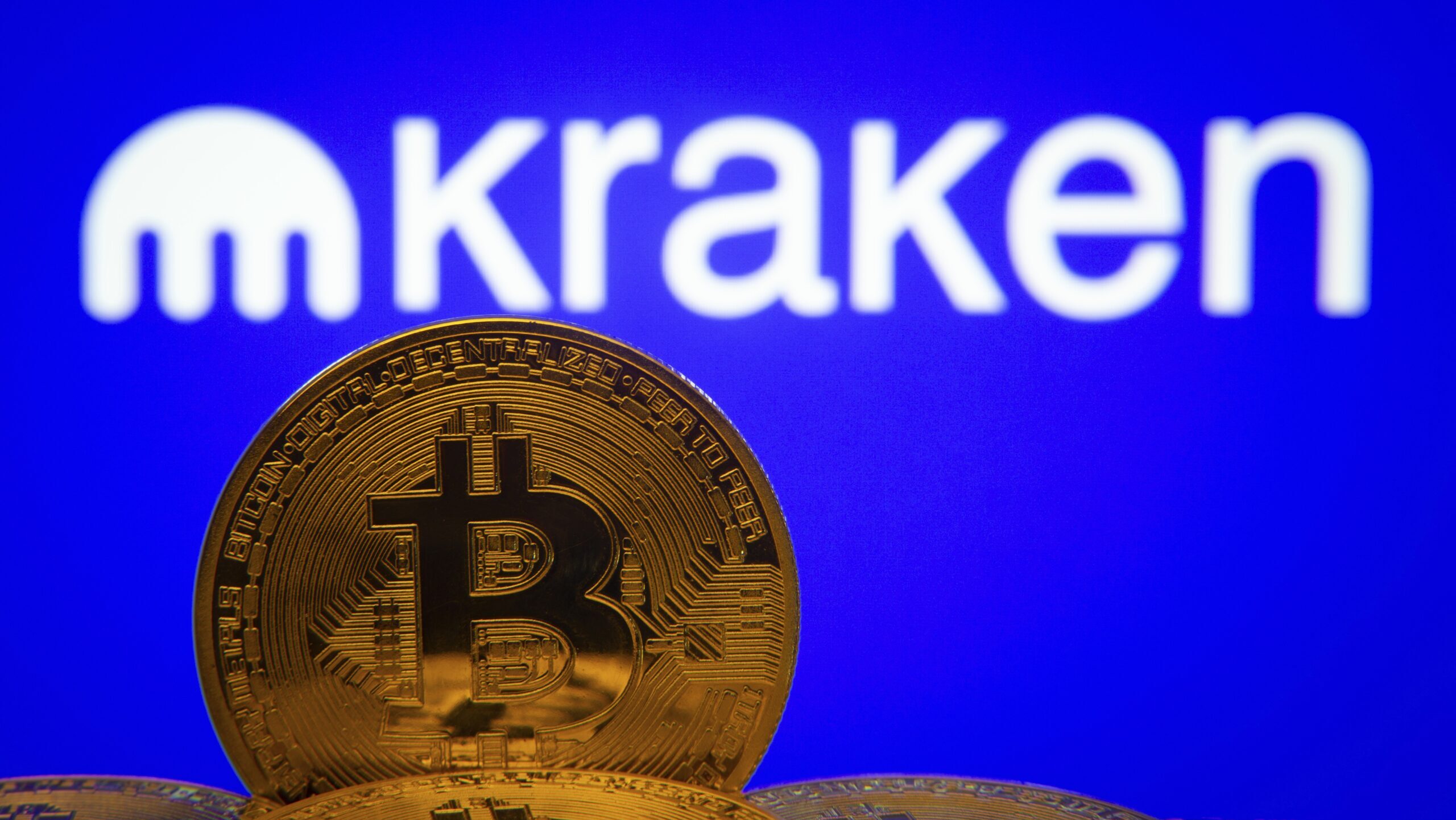 A golden representation of a bitcoin stands in the foreground before the Kraken logo.