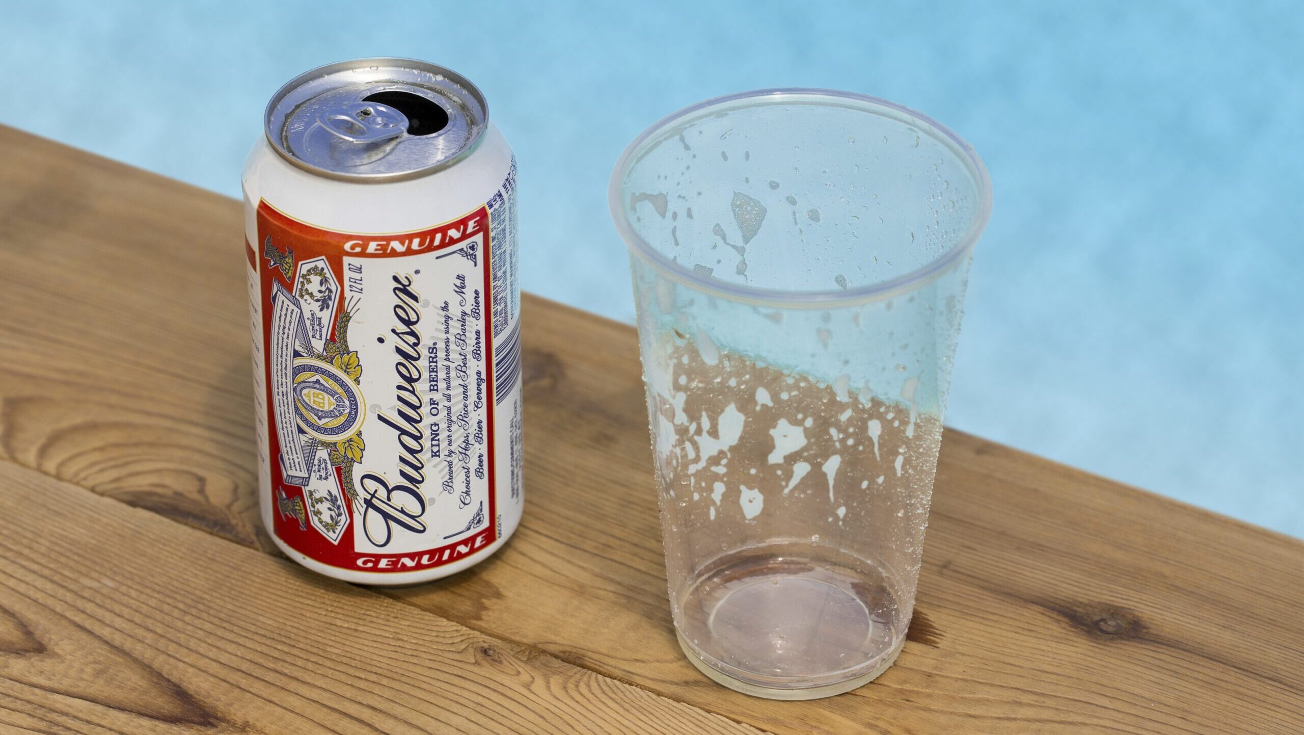 A can of Budweiser beer is shown next to a plastic disposable cup on a wooden table by a swimming pool.