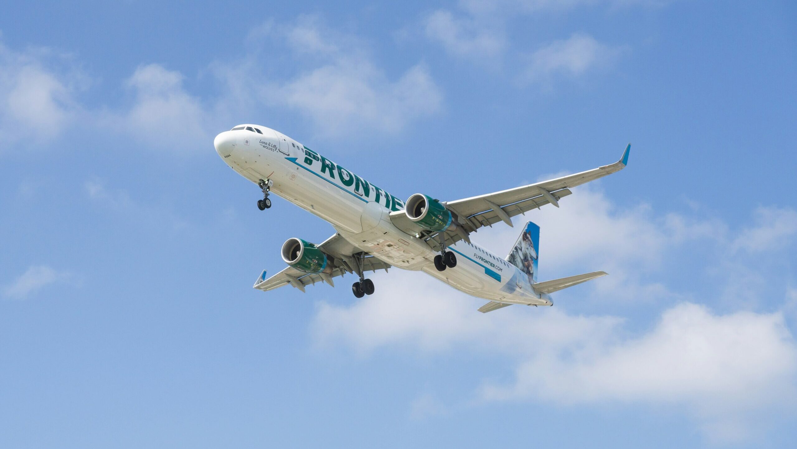 A Frontier Airlines flight is shown landing against a partly cloudy sky.