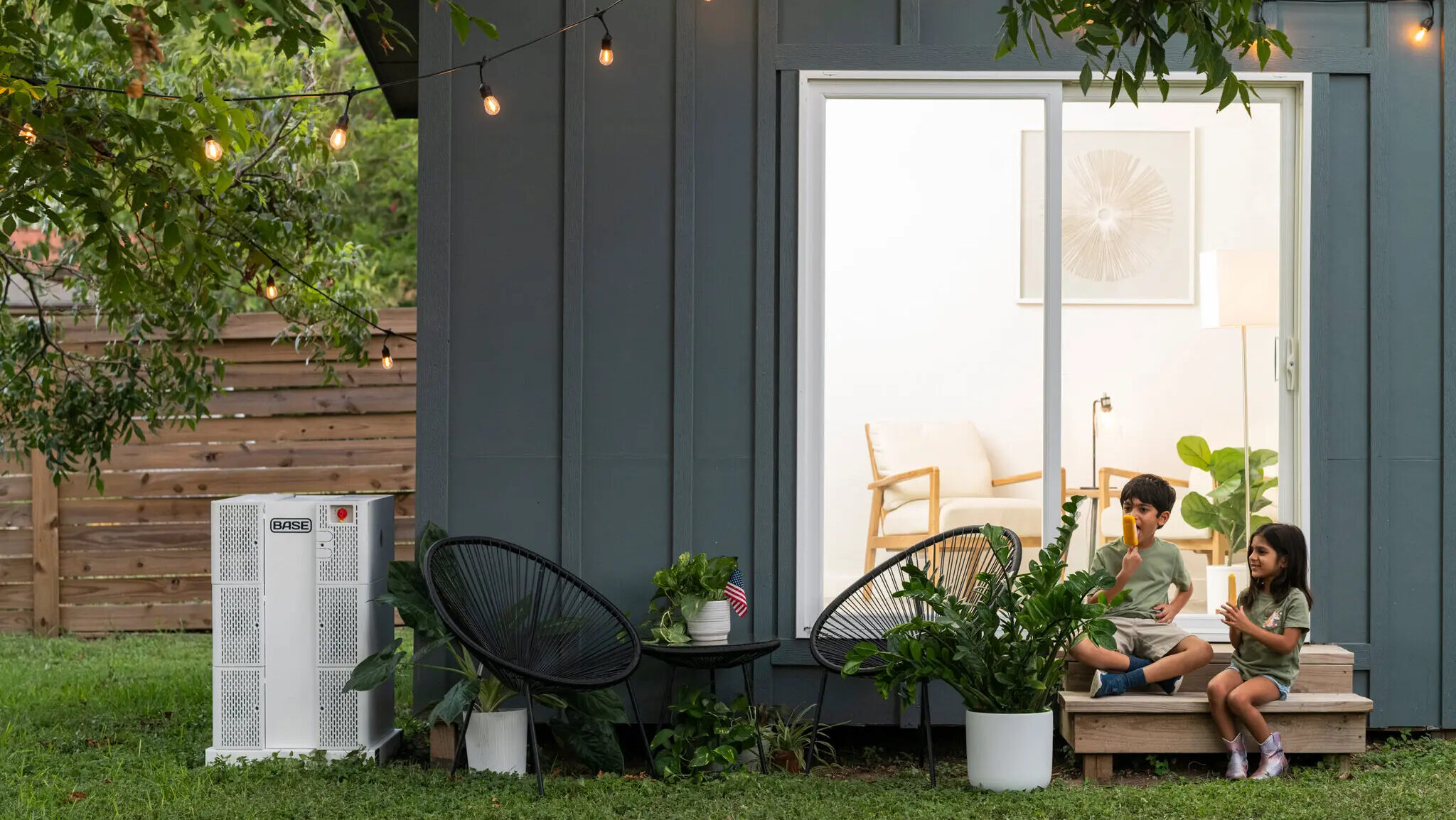 A Base Power home battery is shown next a residence where two children are enjoying frozen popsicles on the doorstep.